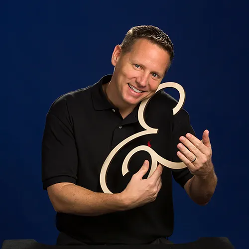 Chuck Norman, owner of S&A Communications, smiles warmly while holding a large wooden ampersand symbol, wearing a black polo shirt with the company logo