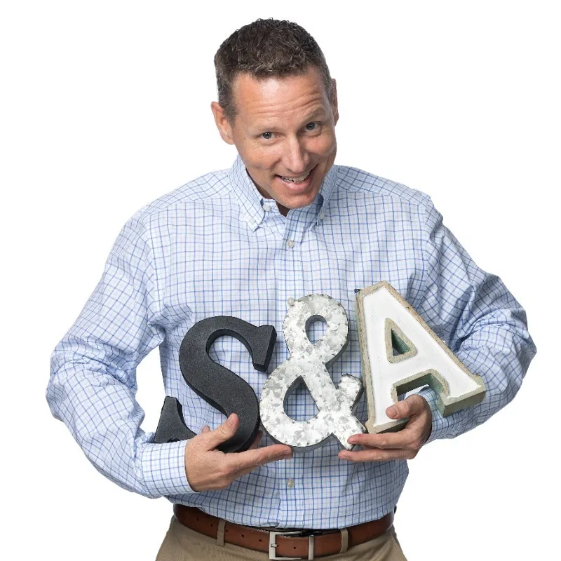 Chuck Norman, owner of S&A Communications, happily holds up decorative 'S', ampersand, and 'A' letters representing his company logo, wearing a blue plaid shirt.