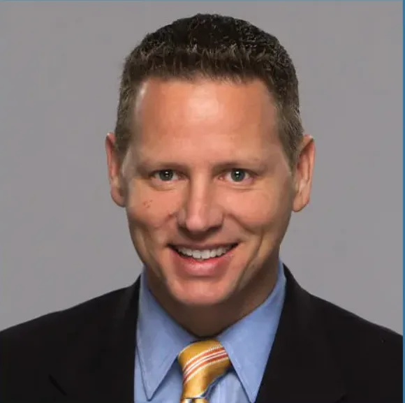 Professional headshot of Chuck Norman smiling confidently in a black suit, light blue shirt, and yellow striped tie against a neutral background