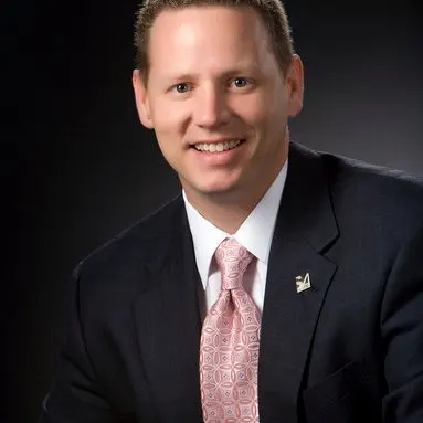 Professional headshot of Chuck Norman in a dark suit and patterned pink tie, smiling confidently against a dark background